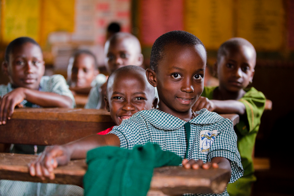 Ugandan student smiles at her desk, surrounded by classmates