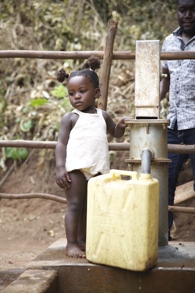 young african girl stands next to the clean water well pumping water into her jerrycan
