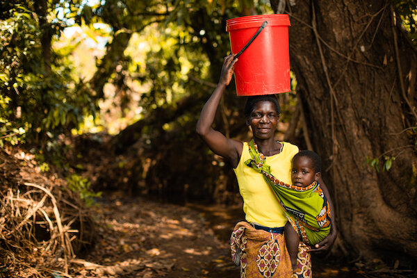 Woman from the chifenthe village holds her baby and stands with a bucket on her head in front of the stream where she used to collect her water before the installation of the new water well in her village